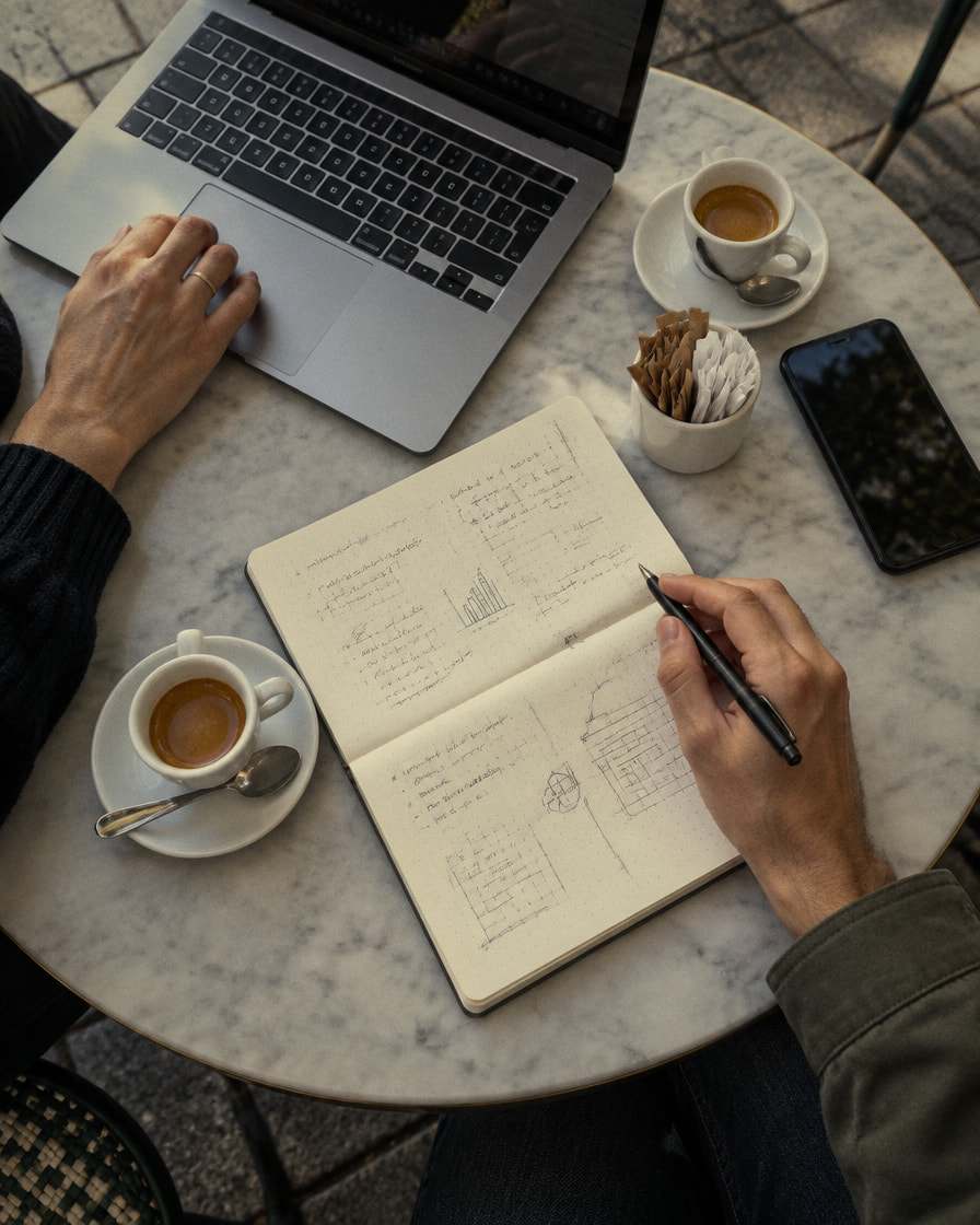 Visto dall’alto, un incontro al bar: su un tavolino di marmo un portatile aperto, un quaderno di appunti e due caffè